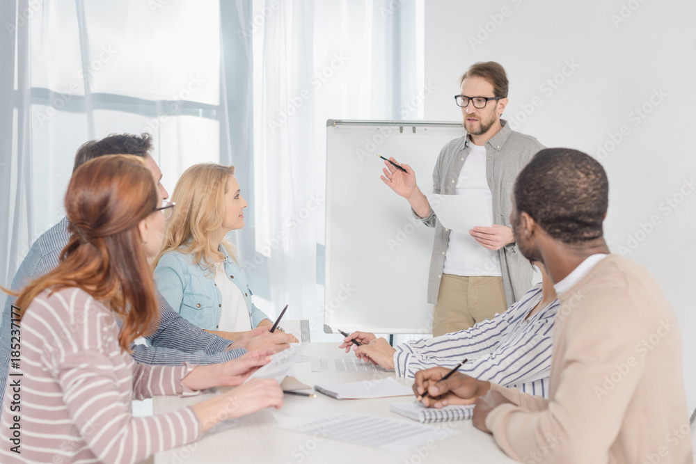 Fototapeta premium middle aged man in in eyeglasses with papers standing at whiteboard and having conversation with his team