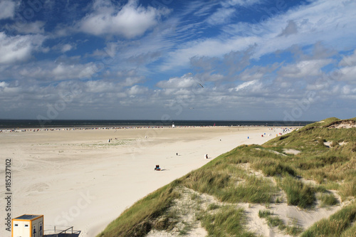 Weitläufiger, gelbweißer, breiter Nordseestrand, mit grün bewachsenen Dünen und wenigen Badegästen mit abwechslungsreicher, vielschichtiger, weißer Bewölkung vor blauem Himmel  photo