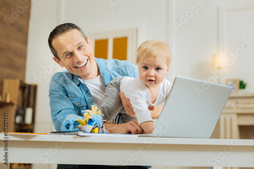 Crawling. Positive excited young father feeling delighted while being at home and seeing his adorable little baby crawling on the table over his modern laptop