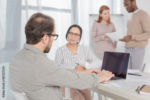 mature man using laptop with blank screen while multiethnic people sitting and standing behind