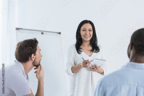 smiling asian psychoanalyst taking notes in notebook while multiethnic men sitting in chairs