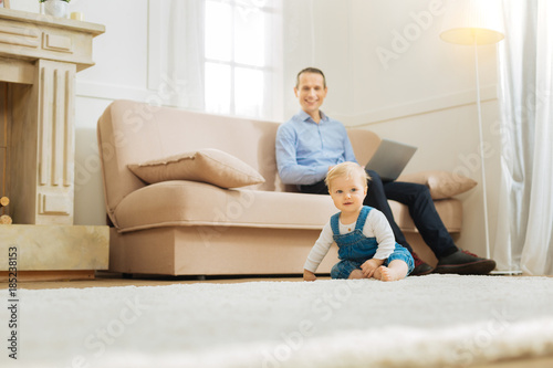 Active morning. Emotional happy young parent looking glad while sitting on a sofa with a laptop in his hands and his cute little baby playing on a soft carpet near him