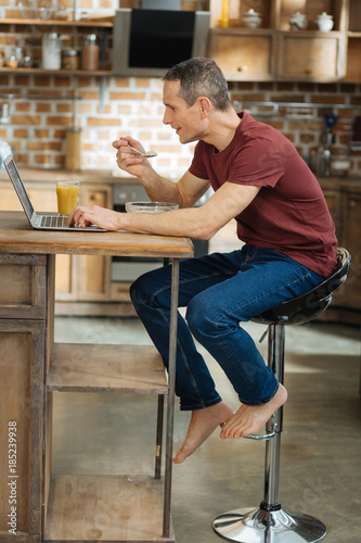 Keep feet. Delighted man sitting at the table and using computer while having breakfast