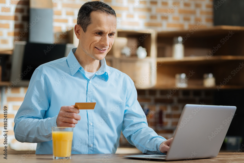 Competent shopper. Cheerful male person keeping smile on his face and holding credit card while looking at his computer Competent shopper. Cheerful male person keeping smile on his face and holding credit card while looking at his computer