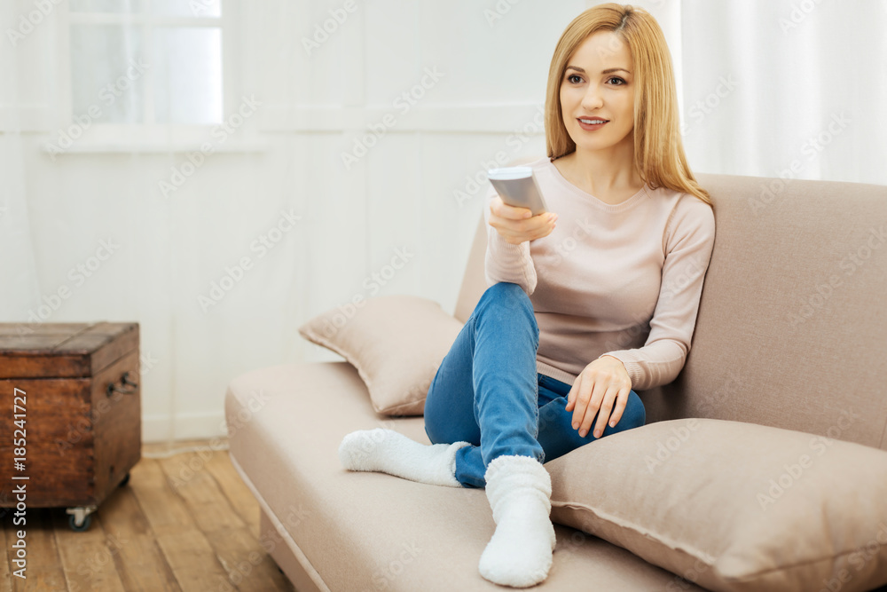 Watching TV. Beautiful smiling young slim blond woman holding a remote control and wearing jeans and a beige sweater and warm socks and sitting on the couch with cushions in the room Watching TV. Beautiful smiling young slim blond woman holding a remote control and wearing jeans and a beige sweater and warm socks and sitting on the couch with cushions in the room