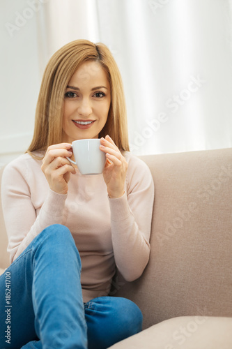 Coffee time. Inspired smiling dark-eyed slim blond woman holding a cup and wearing jeans and a beige sweater and sitting on the couch in the room