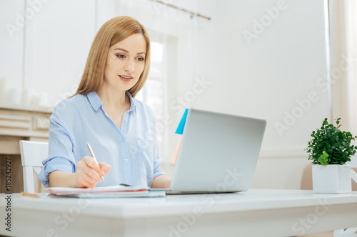 Project. Pretty smiling young blond woman wearing a shirt and writing something down while sitting at the table and working on her laptop and a flower on her table