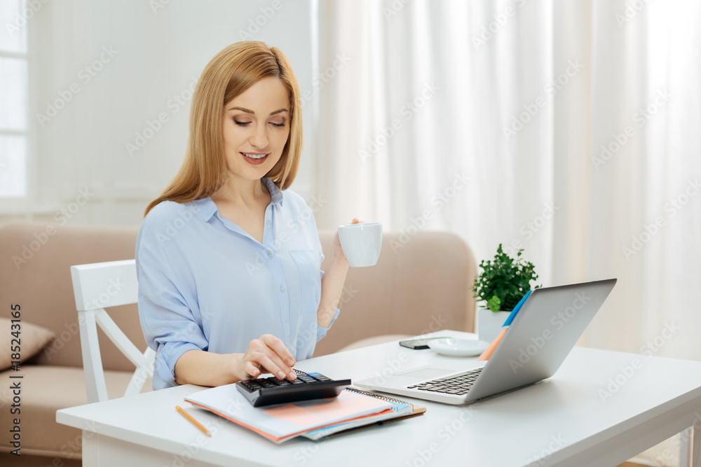 Making calculations. Pretty successful young blond woman smiling and wearing a blue shirt while working on her laptop and using calculator and holding a cup of tea Making calculations. Pretty successful young blond woman smiling and wearing a blue shirt while working on her laptop and using calculator and holding a cup of tea
