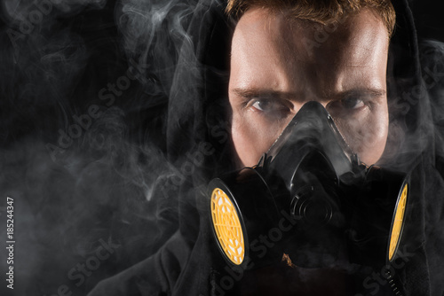 Man in black hood wearing protective filter mask surrounded by clouds of smoke