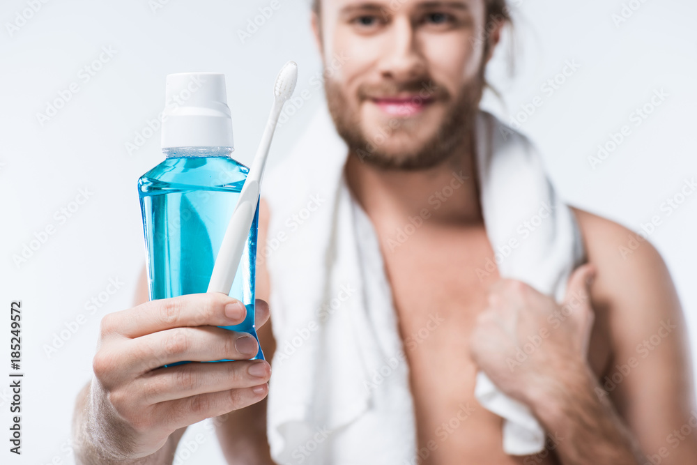 Smiling man with towel around his neck holding tooth rinse and toothbrush in hand, isolated on white Smiling man with towel around his neck holding tooth rinse and toothbrush in hand, isolated on white