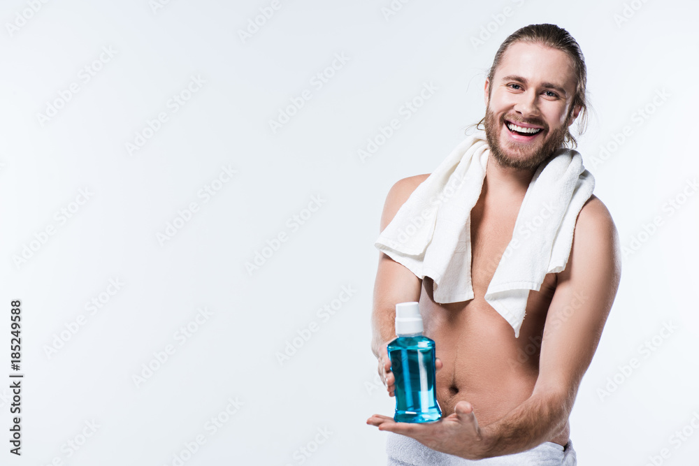 Smiling young man with bath towel around his neck holding bottle with tooth rinse in hands,  isolated on white