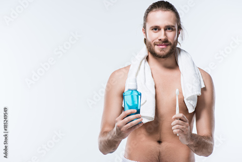 Smiling young man with towel around his neck holding tooth rinse and toothbrush, isolated on white