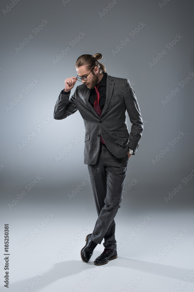 Long haired businessman in glasses looking down while standing with crossed legs, isolated on gray Long haired businessman in glasses looking down while standing with crossed legs, isolated on gray