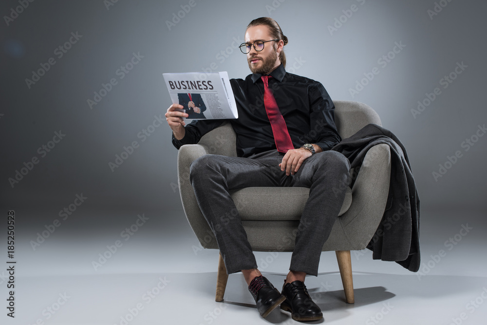 Caucasian bearded businessman in glasses sitting in armchair and reading newspaper, isolated on gray Caucasian bearded businessman in glasses sitting in armchair and reading newspaper, isolated on gray