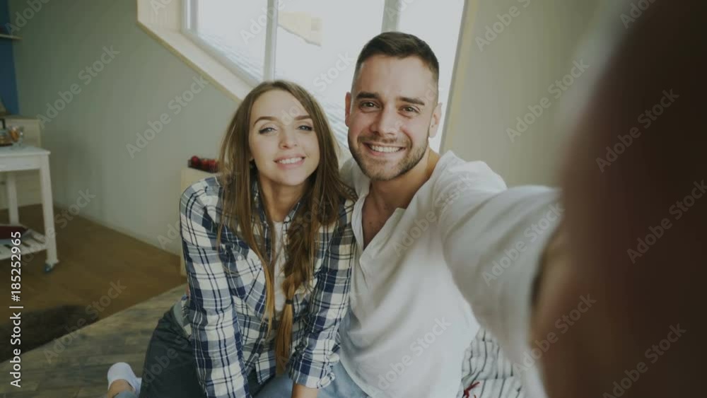 POV of Young smiling and beautiful couple take selfie portrait on smartphone camera and kiss while sitting on bed in the morning