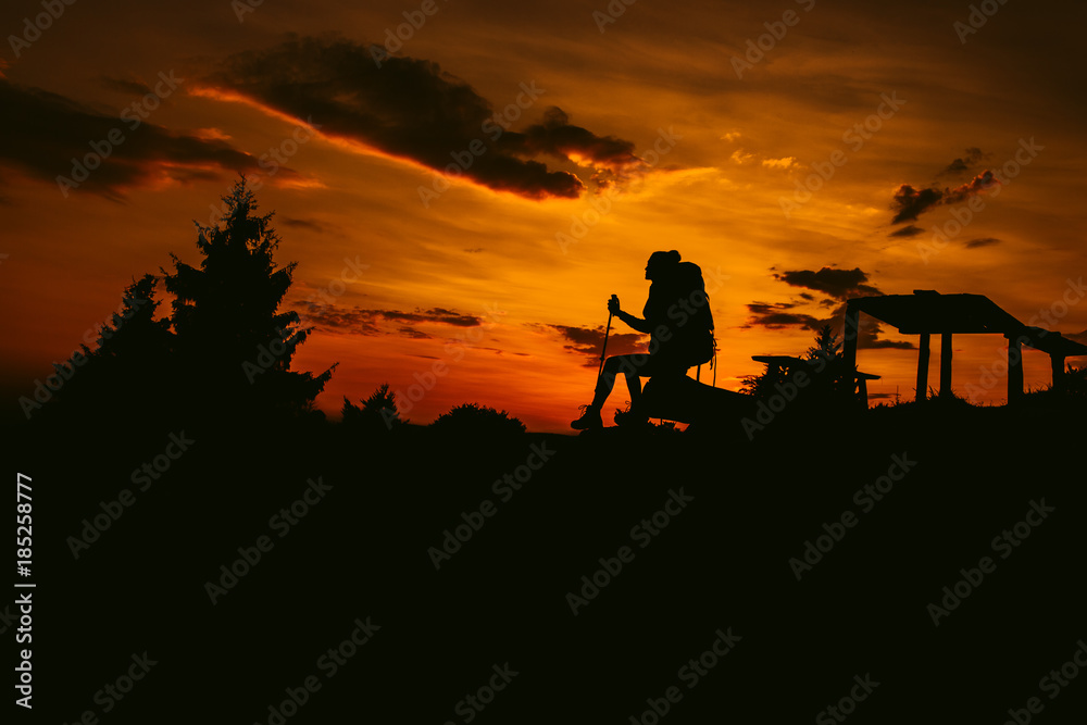 Silhouette hiker woman sit on the bench, with backpack and trekking pole, sunset orange sky on the background. Rest during a hike