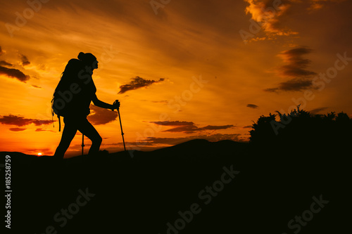 Silhouette sporty woman tracking with backpack and trekking pole, sunset orange sky on the background