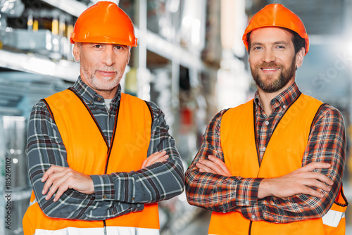 two male workers in safety vests and helmets with crossed arms in storehouse