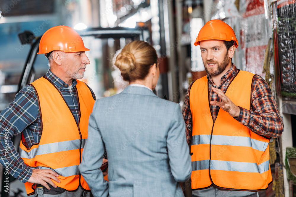 male workers in safety vests and helmets talking with inspector in storehouse male workers in safety vests and helmets talking with inspector in storehouse