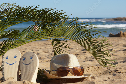 Beach slippers with painted faces and a hat with sunglasses in a tropical resort under a small palm tree