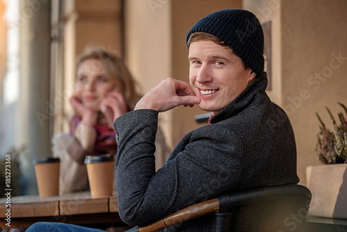 Waist up portrait of young guy sitting at cafe with pretty girl. He is looking at camera while she is talking on phone on the background. They are enjoying hot coffee outside
