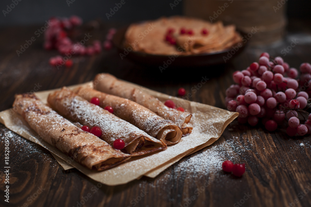 sweet fresh pancakes with a pile and pancakes on baking paper, rolled with tubules, viburnum berries on a dark wooden table on a dark gray background