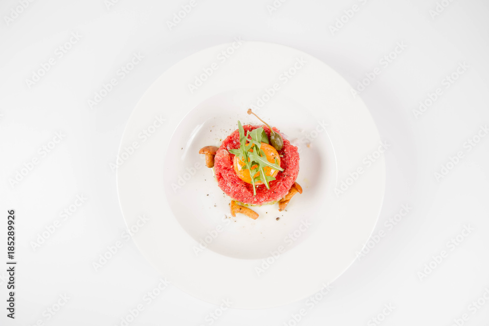 Steak tartare served with egg yolk, onions, caper, mushroom and rocket salad on a white background Steak tartare served with egg yolk, onions, caper, mushroom and rocket salad on a white background