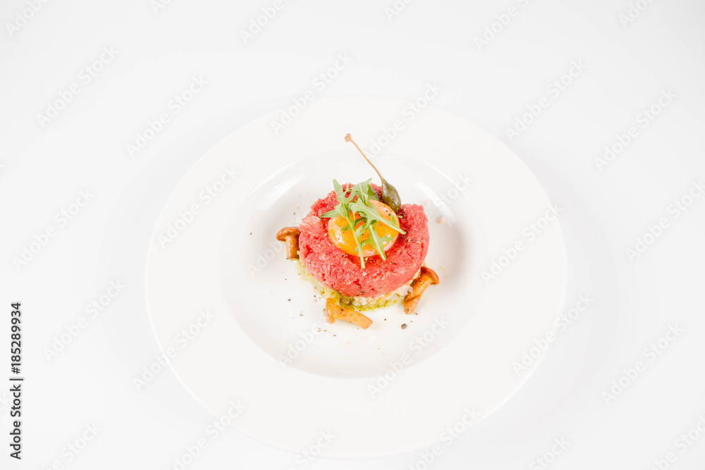 Steak tartare served with egg yolk, onions, caper, mushroom and rocket salad on a white background Steak tartare served with egg yolk, onions, caper, mushroom and rocket salad on a white background