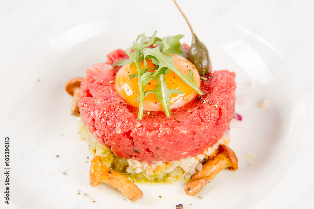 Steak tartare served with egg yolk,  onion, pickle,  caper, mushroom and rocket salad on a white background