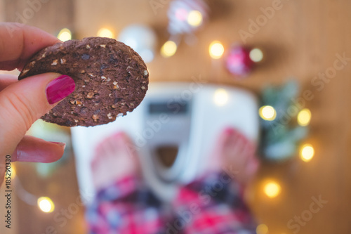 Close up of a female hand holding a chocolate cookie with a blurred background with a weight scale and christmas lights, holiday diet and weight gain concept 