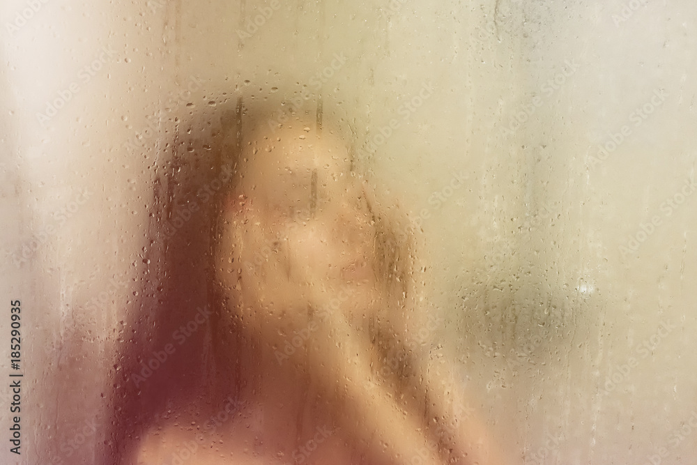Beautiful woman in the shower behind glass with drops. Yellow glow.