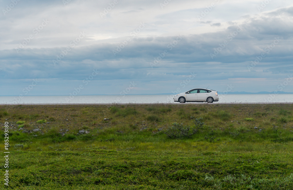White car on a road against a background of green grass, sea and cloudy sky