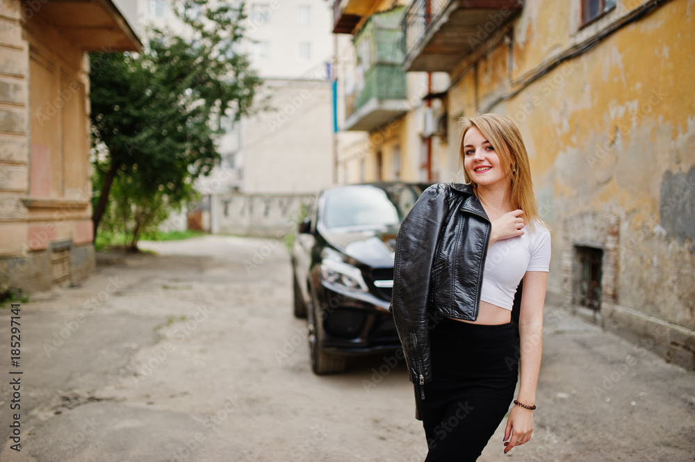 Elegant blonde girl wear on black leather jacket posing at streets of town background luxury car.