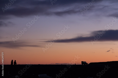 Two people walking across a rocky landscape toward a camper van in silhouette against a colorfuly sky at sunset © Bob