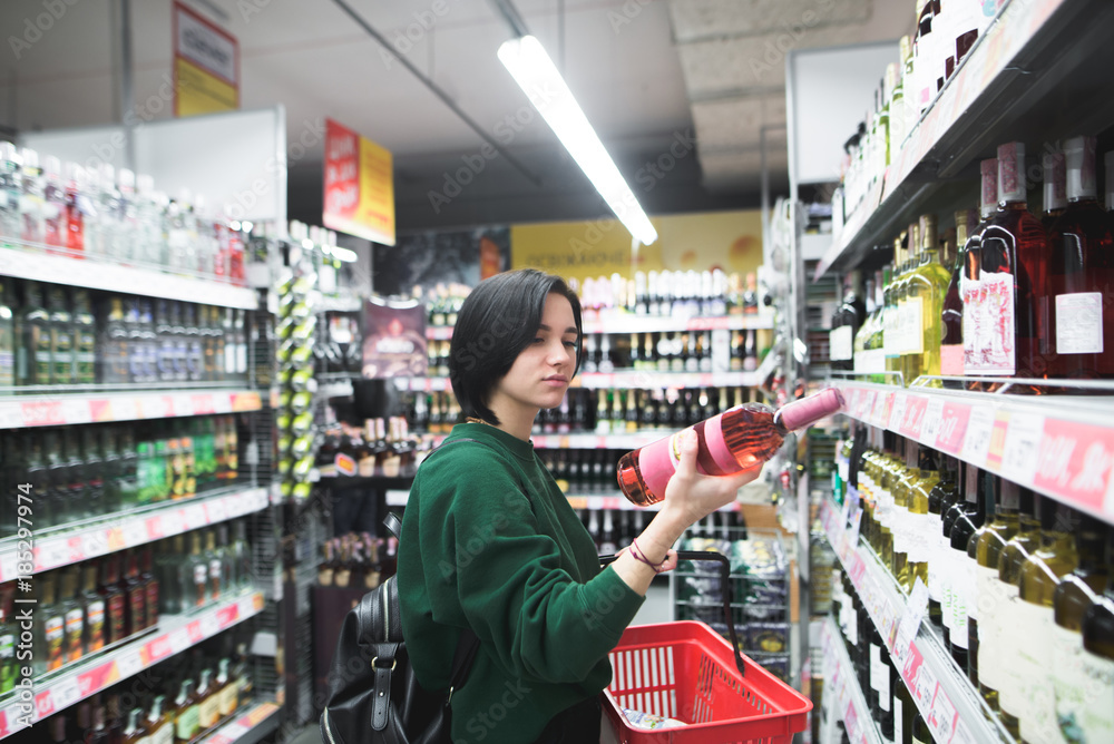 A beautiful girl looks at the wine bottle carefully when shopping at the supermarket. The girl reads a wine label. Shopping in a supermarket.