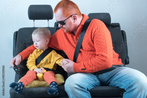 Car seat belt. A happy child is sitting in auto armchair next to man with red hair, beard and mustache in yellow shirt, glasses with laptop. The concept of road safety.