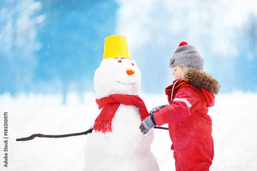 Little boy in red winter clothes having fun with snowman in snowy park
