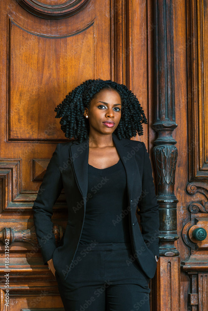 African American Business Woman working in New York. Wearing blazer, v neck  undershirt, young pretty black teacher with braid hairstyle standing by  vintage style classroom doorway, looking at you.. Stock Photo |