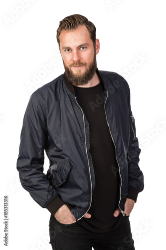 Handsome bearded man standing against a white background wearing a black tshirt and blue jacket in jeans. Looking at camera.