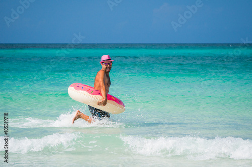 Happy handsome man in pink hat is running with big inflatable tube on the coast of Caribbean Sea in summer sunny day
