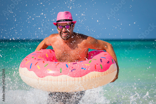 Happy handsome man in pink hat is having fun with big inflatable ring on the coast of Caribbean Sea in summer sunny day