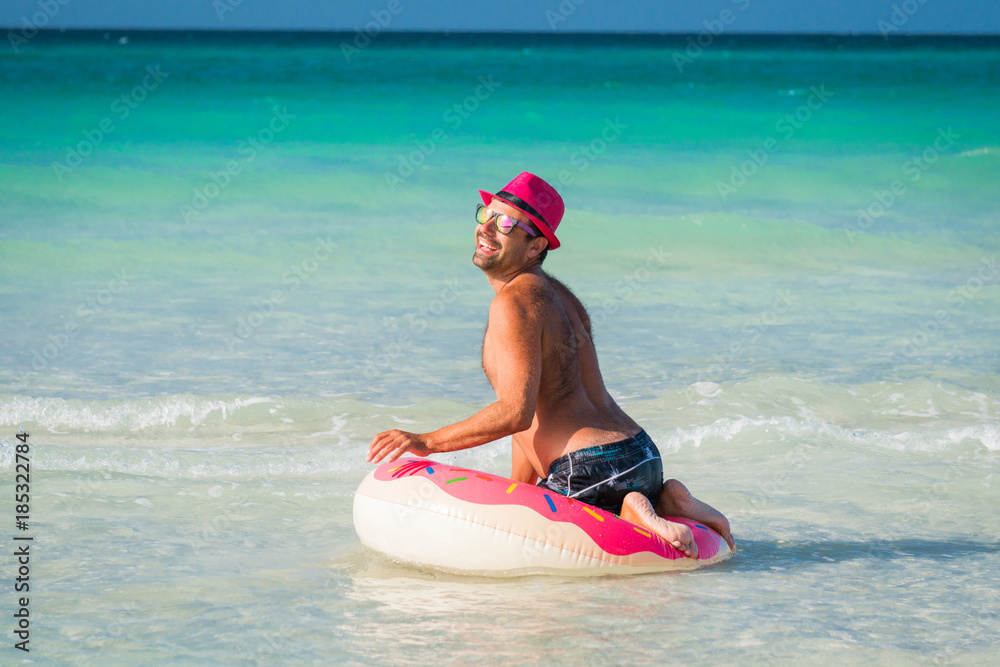 Happy handsome smiling man in pink hat has fun with big inflatable tube on the coast of Caribbean Sea in summer sunny day