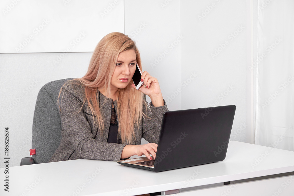 Close up of an interested businesswoman in a suit working at her white office. She is talking on phone and work with laptop Close up of an interested businesswoman in a suit working at her white office. She is talking on phone and work with laptop