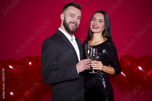 Stylish beautiful smiling couple of lovers with glasses of champagne on a red background with balls in the shape of heart. Women's Day or March 8th. Valentine's Day