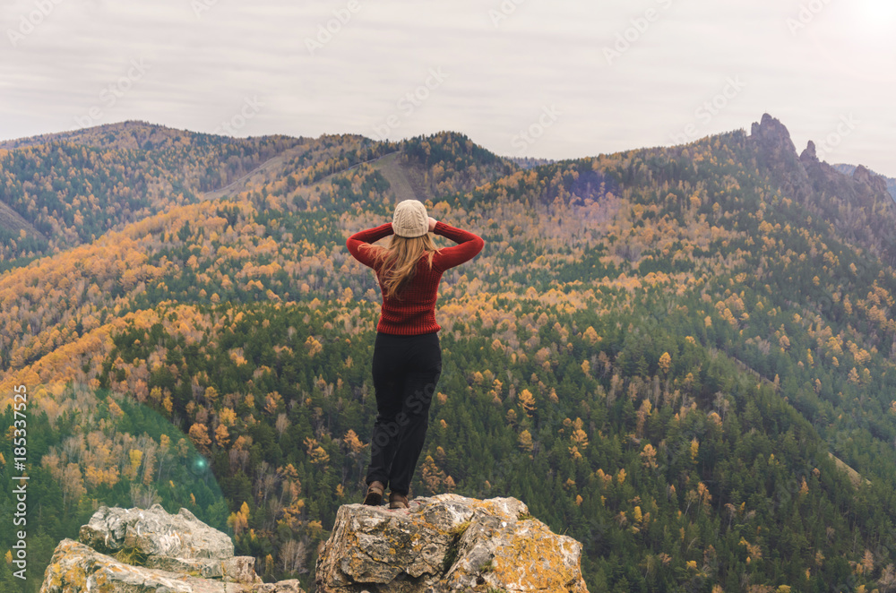 Obraz premium A girl in a red jacket looks out into the distance on a mountain, a view of the mountains and an autumnal forest by an overcast day. Free space for text