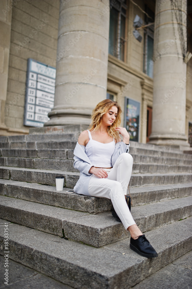 Stylish curly blonde model girl wear on white with cup of coffee posing on stairs outdoor. Stylish curly blonde model girl wear on white with cup of coffee posing on stairs outdoor.