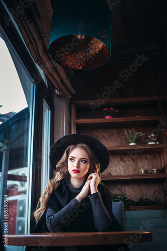 Young beautiful fashionable lady sitting near big window in cafe and holding cup of coffee. Attractive woman wearing stylish black hat. © selenit