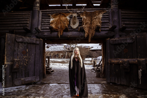 Outdoors portrait of beautiful furious scandinavian warrior ginger woman in a traditional clothes with fur collar, with sword in her hand and wooden Viking Village view on the background.