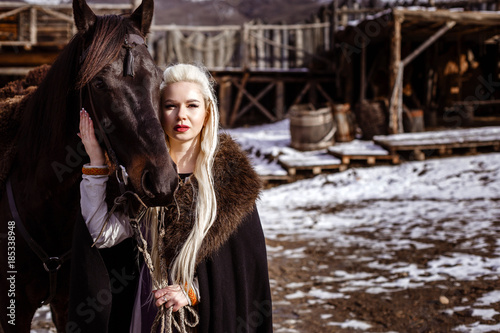 Bild auf Leinwand Outdoors portrait of beautiful furious scandinavian warrior ginger woman in a traditional clothes with fur collar, with sword in her hand and wooden Viking Village view on the background