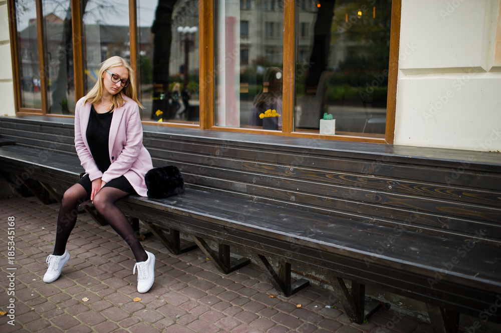 Blonde girl at glasses and pink coat, black tunic sitting on bench at street. Blonde girl at glasses and pink coat, black tunic sitting on bench at street.